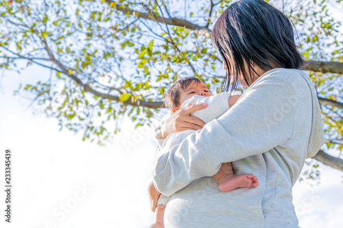 木洩れ日のある樹木の下で赤ちゃんを抱く妊娠後期の幸せなお母さん。育児、妊娠、家族、親子、愛、幸せイメージ