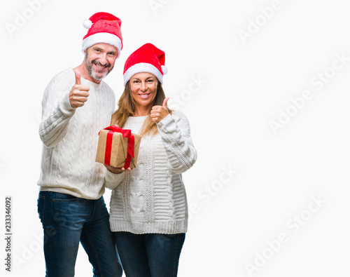 Middle age hispanic couple wearing christmas hat and holding gift over isolated background happy with big smile doing ok sign, thumb up with fingers, excellent sign