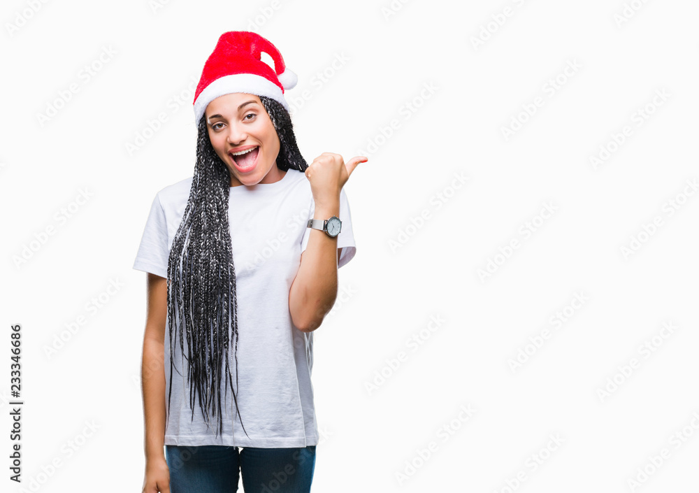 Young braided hair african american girl wearing christmas hat over isolated background smiling with happy face looking and pointing to the side with thumb up. Young braided hair african american girl wearing christmas hat over isolated background smiling with happy face looking and pointing to the side with thumb up.