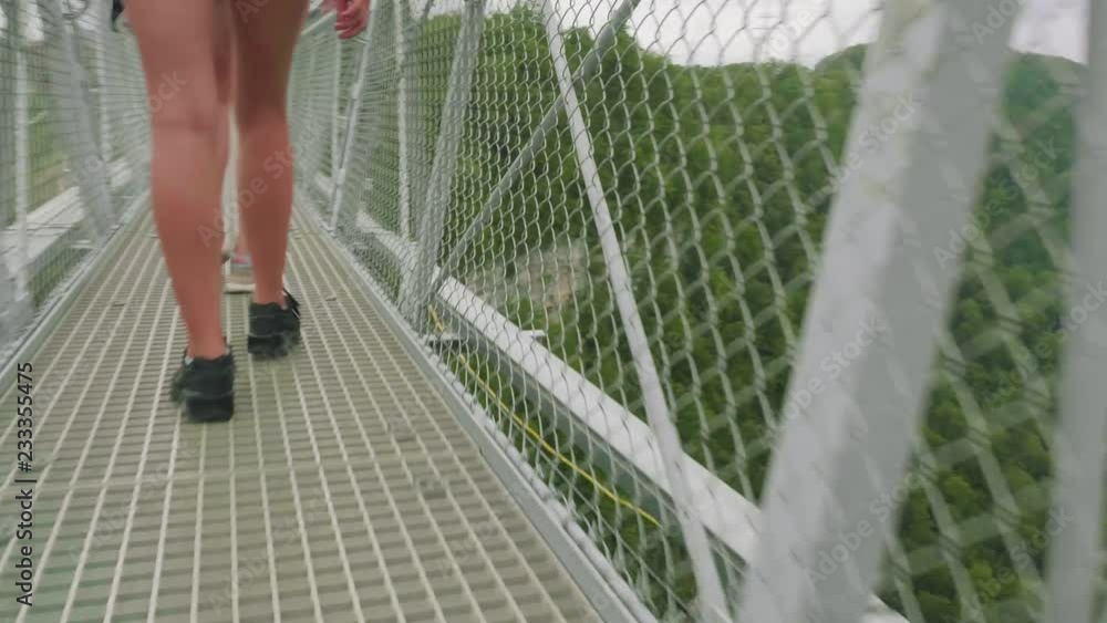 Close-up view of white female sport legs in athletic shoes reaching across suspended lattice bridge high in mountains Close-up view of white female sport legs in athletic shoes reaching across suspended lattice bridge high in mountains