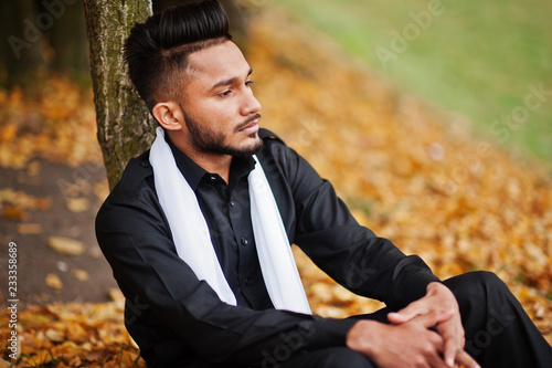 Indian stylish man in black traditional clothes with white scarf posed outdoor against yellow autumn leaves tree.