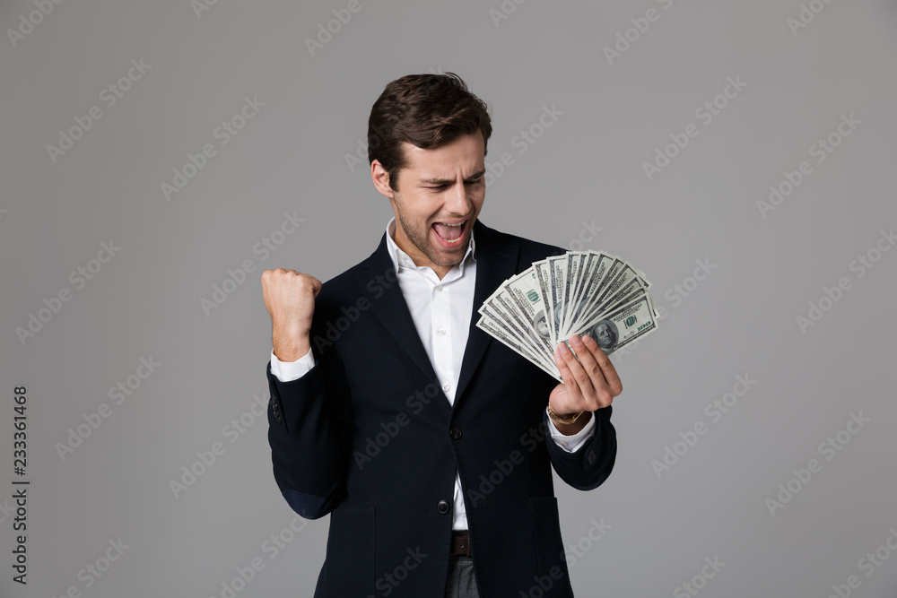 Image of ecstatic businessman 30s in suit smiling and holding fan of money banknotes, isolated over gray background Image of ecstatic businessman 30s in suit smiling and holding fan of money banknotes, isolated over gray background