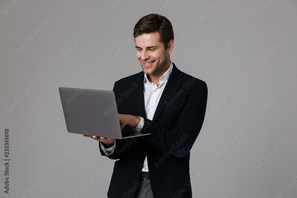 Image of pleased businessman 30s in suit rejoicing while using laptop, isolated over gray background