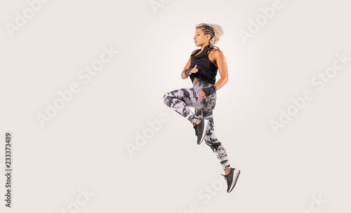 Beautiful female Middle Eastern fitness athlete with modern funky hairstyle and wearing sports clothing doing a dramatic leap in studio with white background