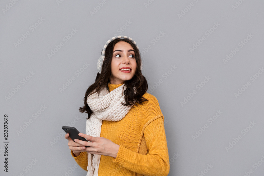 Portrait of brunette woman wearing ear muffs and scarf using mobile phone, isolated over gray background Portrait of brunette woman wearing ear muffs and scarf using mobile phone, isolated over gray background
