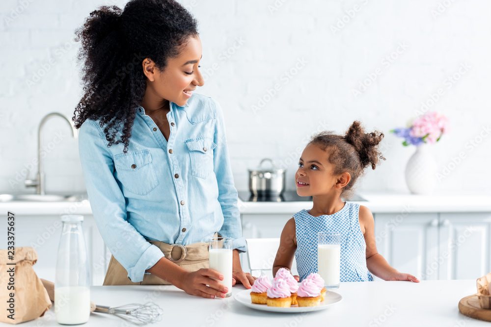 african american mother and daughter looking at each other in kitchen, milk and cupcakes on table african american mother and daughter looking at each other in kitchen, milk and cupcakes on table