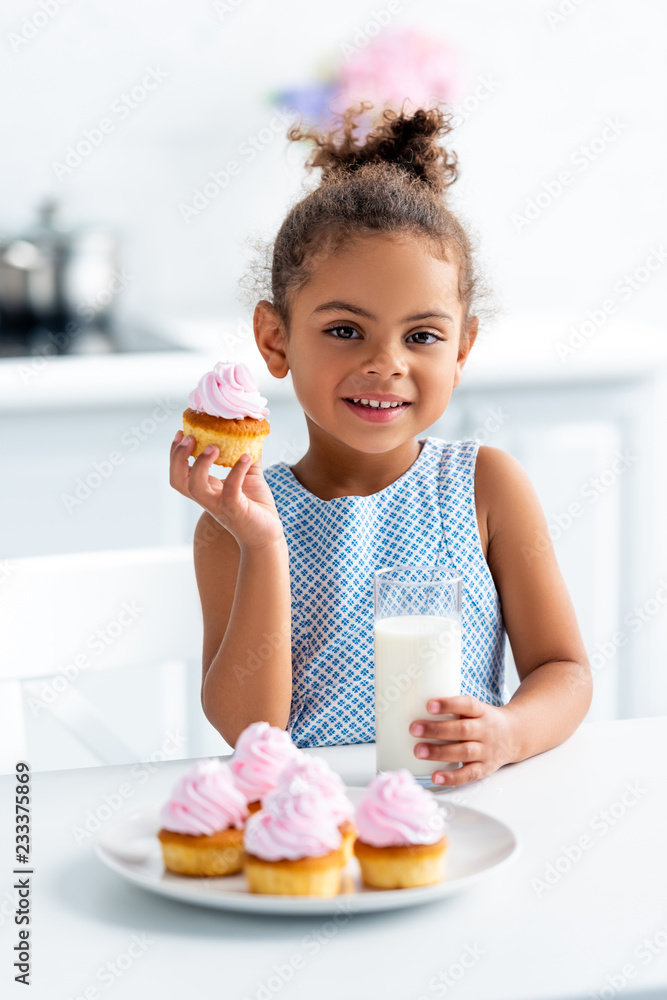 adorable african american kid holding cupcake and glass of milk in kitchen, looking at camera adorable african american kid holding cupcake and glass of milk in kitchen, looking at camera