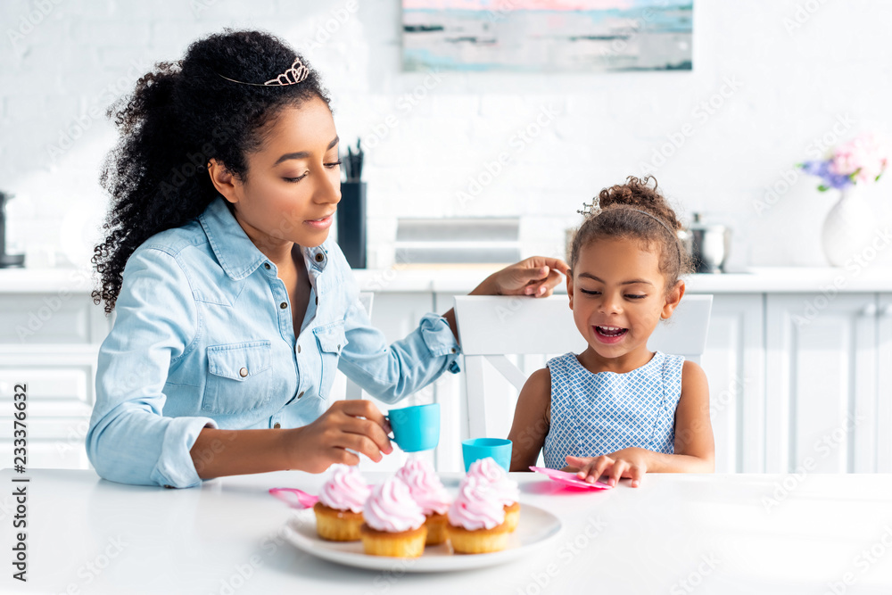 african american mother and daughter with tiaras sitting at table with muffins in kitchen african american mother and daughter with tiaras sitting at table with muffins in kitchen