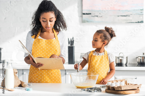 african american mother reading cookbook and daughter preparing dough in kitchen photo