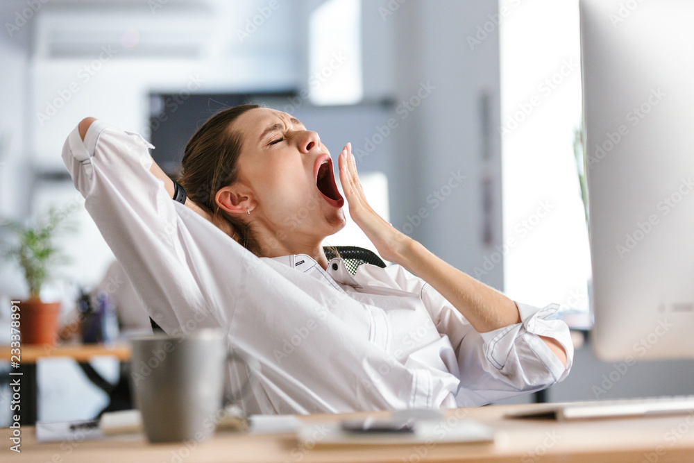 Sleepy young woman dressed in shirt sitting at her workplace - obrazy, fototapety, plakaty 
