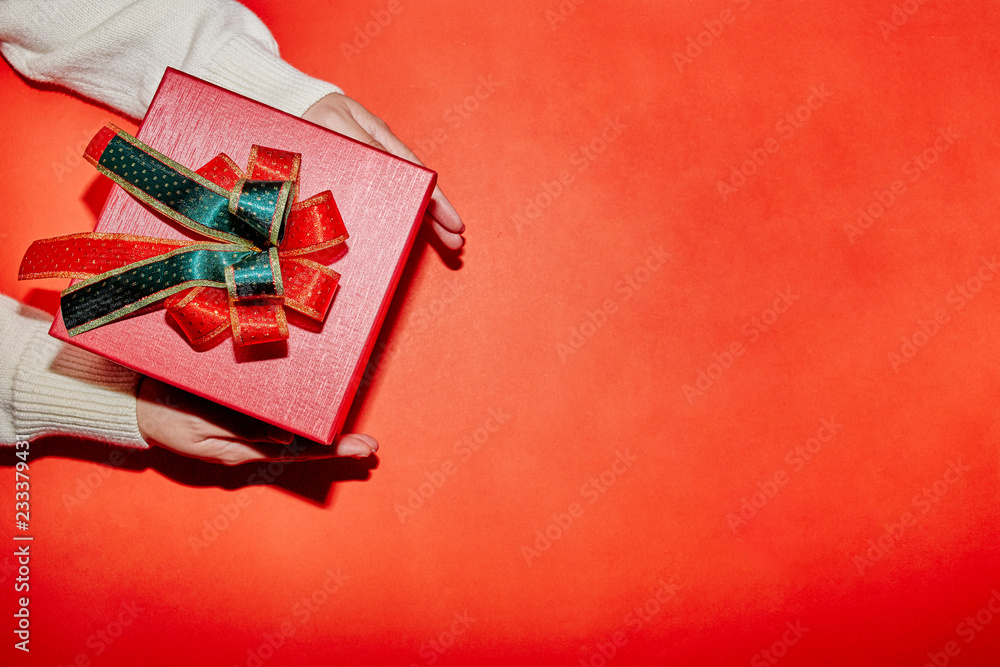 Fototapeta premium female hand in sweater holding a red present box for Christmas, New Year, Valentine'a day on red background, top view with copy space