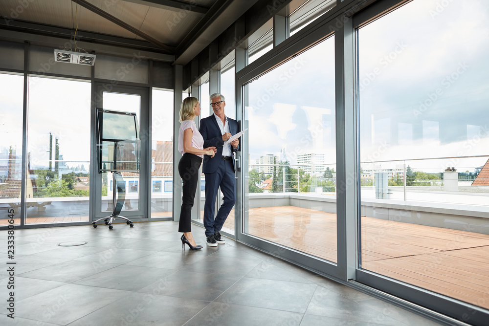 Businessman and woman standing in office, discussing project, holding documents Businessman and woman standing in office, discussing project, holding documents