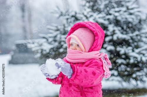 Baby in winter overalls wrapped in a scarf walks on a snow-covered street and sculpts from snow.
