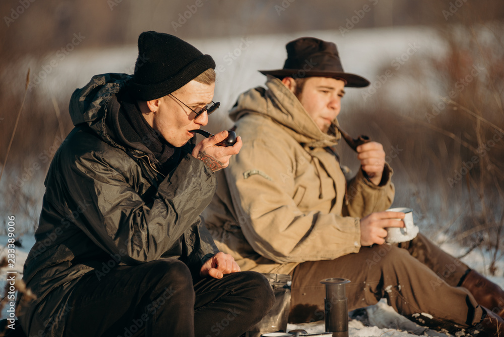 Men is smoking tobacco-pipe and drink coffee. Outdoor portrait of hikker in black sunglasses sitting outdoors during sunny winter day.