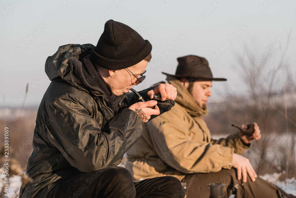 Men is smoking tobacco-pipe and drink coffee. Outdoor portrait of hikker in black sunglasses sitting outdoors during sunny winter day. Men is smoking tobacco-pipe and drink coffee. Outdoor portrait of hikker in black sunglasses sitting outdoors during sunny winter day.