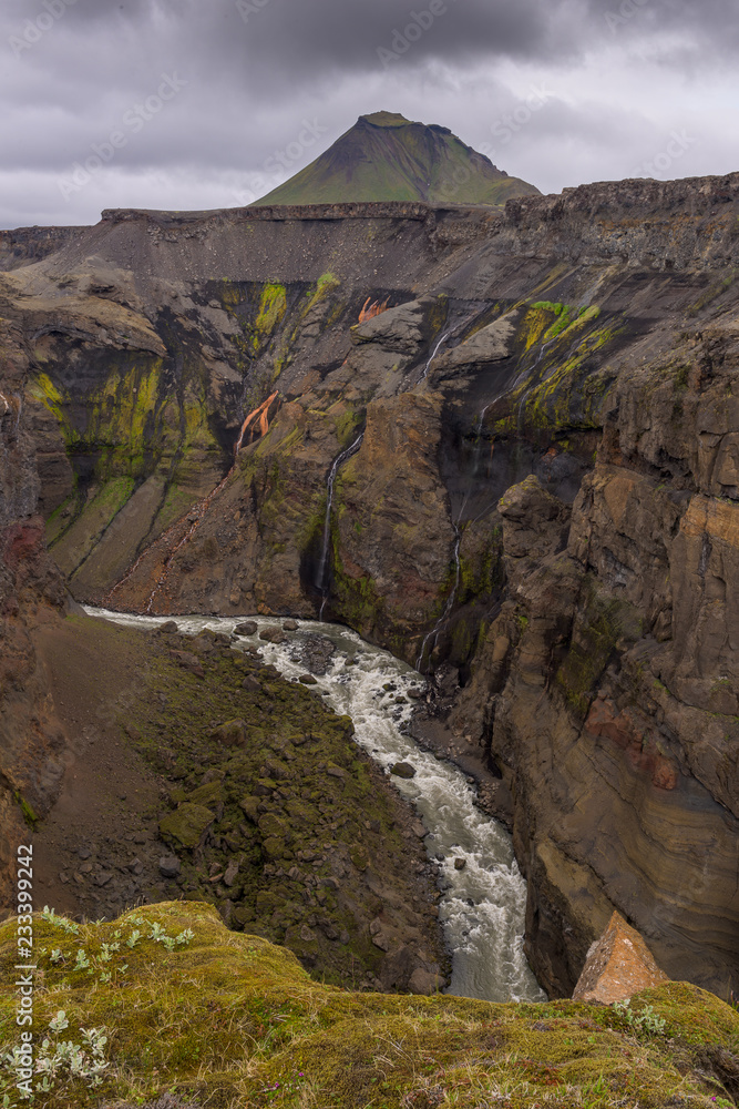 Markarfljótsgljúfur is absolutely incredible place in the middle of Iceland. In the foreground is deep valey with flowing river. We can see geological layers on the cliff. Sky is dark and mysteriuos.