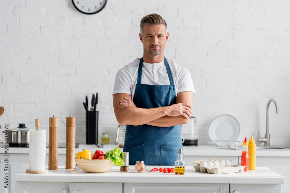 handsome man in apron with arms crossed standing at kitchen handsome man in apron with arms crossed standing at kitchen