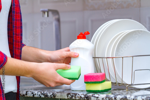 Different clean plates in dish drying rack, dish sponges and dishwashing detergent on the table on kitchen counter.