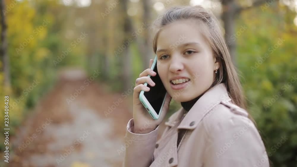 Attractive Girl teenager n with a book talking on mobile phone while walking in beautiful autumn park. Happy Girl teenager talking on mobile phone, outdoors Attractive Girl teenager n with a book talking on mobile phone while walking in beautiful autumn park. Happy Girl teenager talking on mobile phone, outdoors