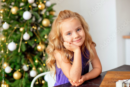 A girl with blond curly hair sits at a table, in the background a Christmas tree, dreams of gifts. Christmas concept, new year. photo