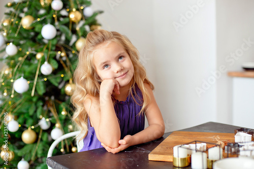 A girl with blond curly hair sits at a table, in the background a Christmas tree, dreams of gifts. Christmas concept, new year. photo