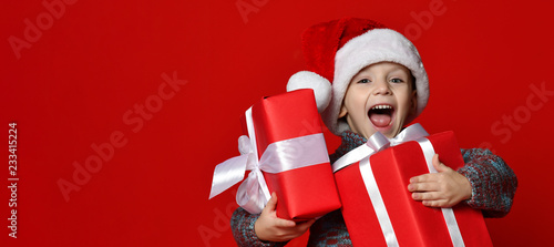 Funny smiling child in Santa red hat holding Christmas gift in hand.