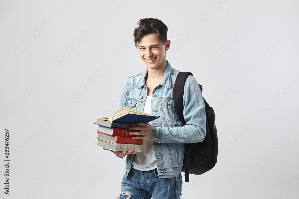 Smiling young dark-haired guy with black backpack on his shoulder dressed in a white t-shirt, jeans and a denim jacket holds books in his hands on the white background in the studio Smiling young dark-haired guy with black backpack on his shoulder dressed in a white t-shirt, jeans and a denim jacket holds books in his hands on the white background in the studio