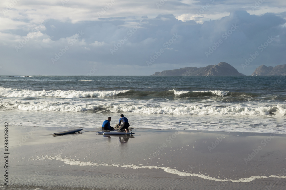 two surfers sitting on the shore of the beach in front of the waves