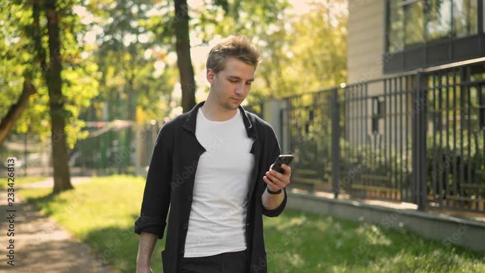 Side view of young man wearing black jacket walking in the park and texting with his smartphone Tracking slow motion medium shot Side view of young man wearing black jacket walking in the park and texting with his smartphone Tracking slow motion medium shot