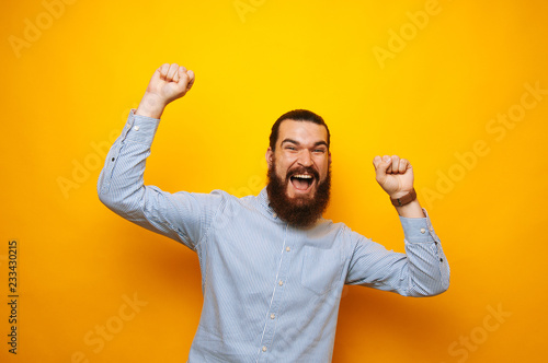 Cheerful bearded man in casual celebrating success over yellow background