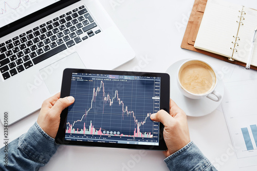 Young trader is sitting in home at table, working on tablet with graphs,diagrams on monitor. Crypto currencies, trader using tablet and computers photo