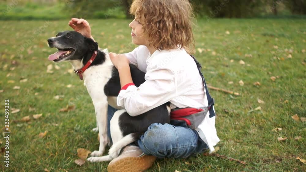 Close portrait of a curly schoolboy with a dog. A boy hugs and strokes his homemade black and white dog sitting on the grass in the park Close portrait of a curly schoolboy with a dog. A boy hugs and strokes his homemade black and white dog sitting on the grass in the park