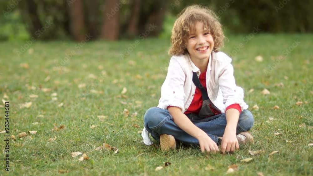 A smiling boy sits on the green grass in the summer park. A schoolboy on vacation, wearing a white shirt, a red T-shirt and jeans, the boy has curly hair A smiling boy sits on the green grass in the summer park. A schoolboy on vacation, wearing a white shirt, a red T-shirt and jeans, the boy has curly hair