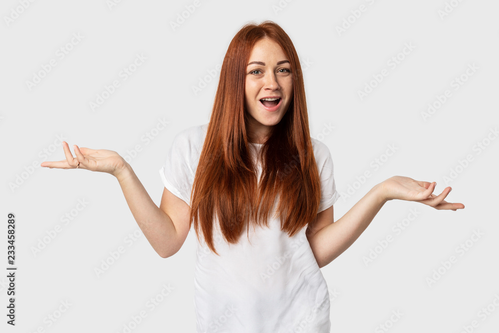 Young emotional European female isolated on grey background. Red-haired girl smiles in a white T-shirt. Shows surprise with hand gestures Young emotional European female isolated on grey background. Red-haired girl smiles in a white T-shirt. Shows surprise with hand gestures