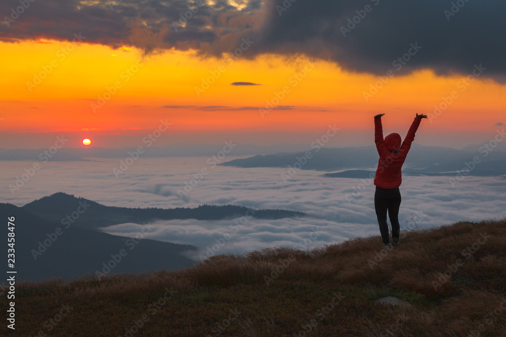 A girl in a red jacket with a hood watching from the top of the mountain sunrise over a mist-covered valley A girl in a red jacket with a hood watching from the top of the mountain sunrise over a mist-covered valley