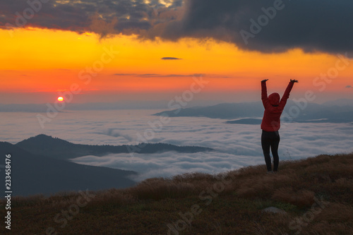 A girl in a red jacket with a hood watching from the top of the mountain sunrise over a mist-covered valley
