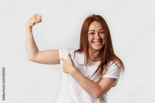 Portrait smiling woman in white t-shirt , isolated on gray background. Showing biceps muscle having lifted arm with clenched fist photo