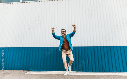 Jumping and smiling. Emotional stylish young man jumping against the colorful wall and putting both hands up