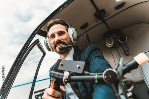 Attractive young man elegant pilot wearing big headphones and smiling while sitting in the helicopter cabin