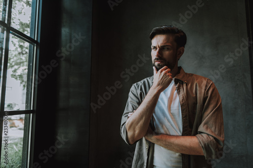 Portrait of pensive unshaven male touching chin with hand while looking at street in apartment