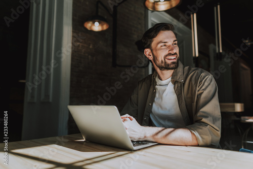 Fotografie Portrait of beaming man with attractive smile working with laptop while locating at table