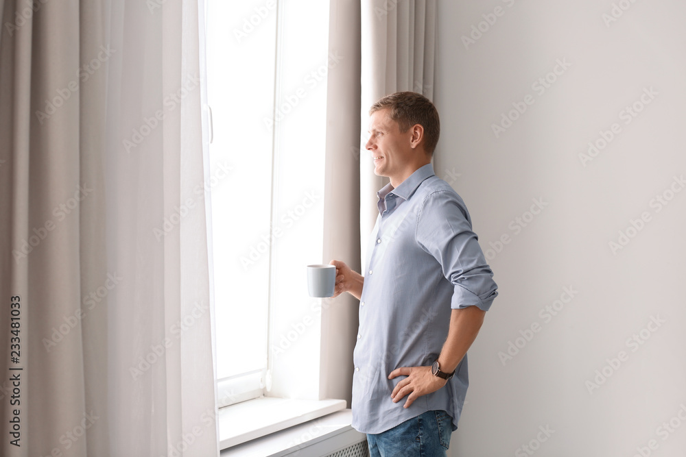 Mature man with cup of drink near window with open curtains at home. Space for text Mature man with cup of drink near window with open curtains at home. Space for text