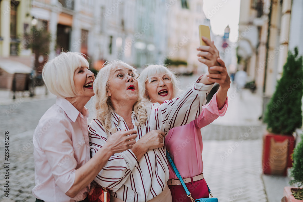 Obraz premium Happy stylish long-haired woman holding phone and making photo with hers friend outdoor