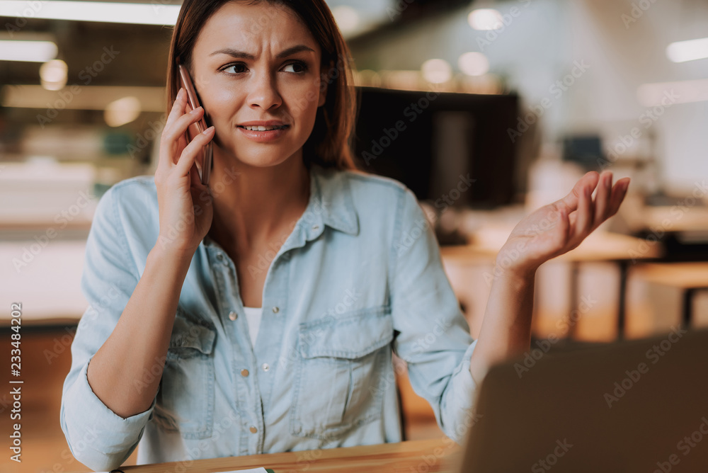 Portrait of attractive woman having phone conversation while sitting at table. She is looking away with confused expression Portrait of attractive woman having phone conversation while sitting at table. She is looking away with confused expression
