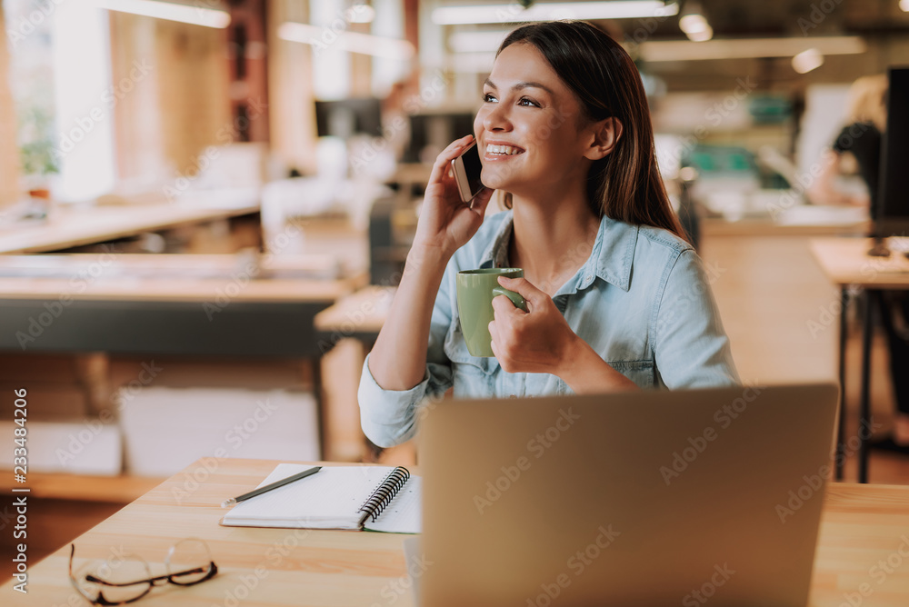 Portrait of charming girl holding cup of coffee and having phone conversation while sitting at office desk with laptop, notebook and pencil. She is looking away and smiling Portrait of charming girl holding cup of coffee and having phone conversation while sitting at office desk with laptop, notebook and pencil. She is looking away and smiling