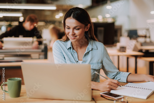 Portrait of beautiful smiling girl working with notebook and holding pencil while sitting at desk with cellphone and cup of coffee