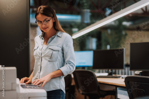 Waist up portrait of charming woman standing near photocopier and smiling. Desk with computers on blurred background
