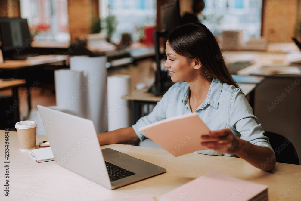 Working process. Portrait of charming woman sitting at office desk with laptop and looking at smartphone with smile Working process. Portrait of charming woman sitting at office desk with laptop and looking at smartphone with smile