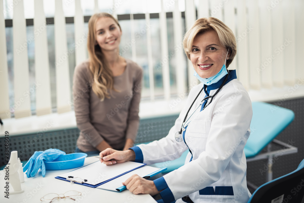 Fototapeta premium Concept of positive professional consultation in therapist system. Waist up portrait of smiling female doctor and young woman sitting in medical office