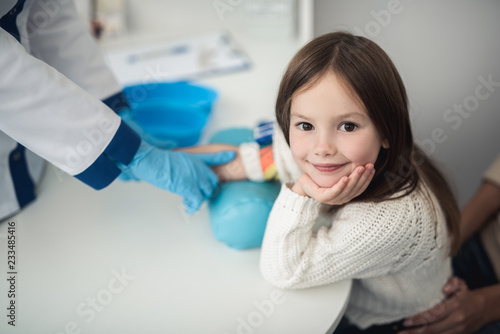 Professional laboratory investigations in healthcare system. Top view portrait of smiling female child ready to have haemanalysis by doctor in medical office